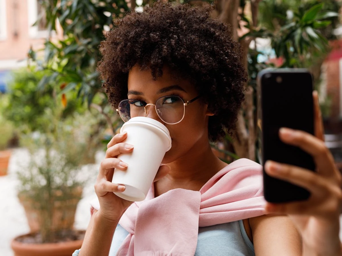 Content creator in glasses sipping takeaway coffee while filming on her smartphone at an outdoor cafe