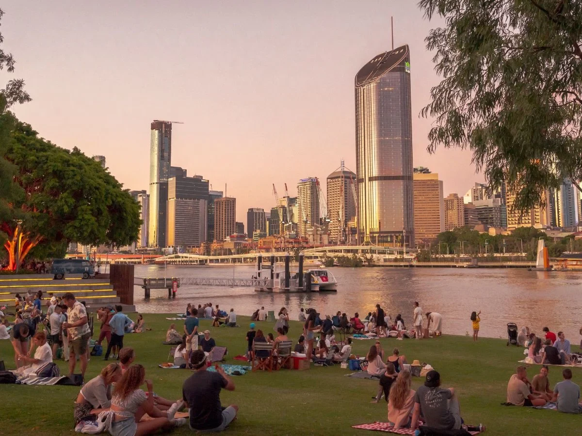 Crowd relaxing on the riverside lawn at sunset with the Brisbane skyline behind