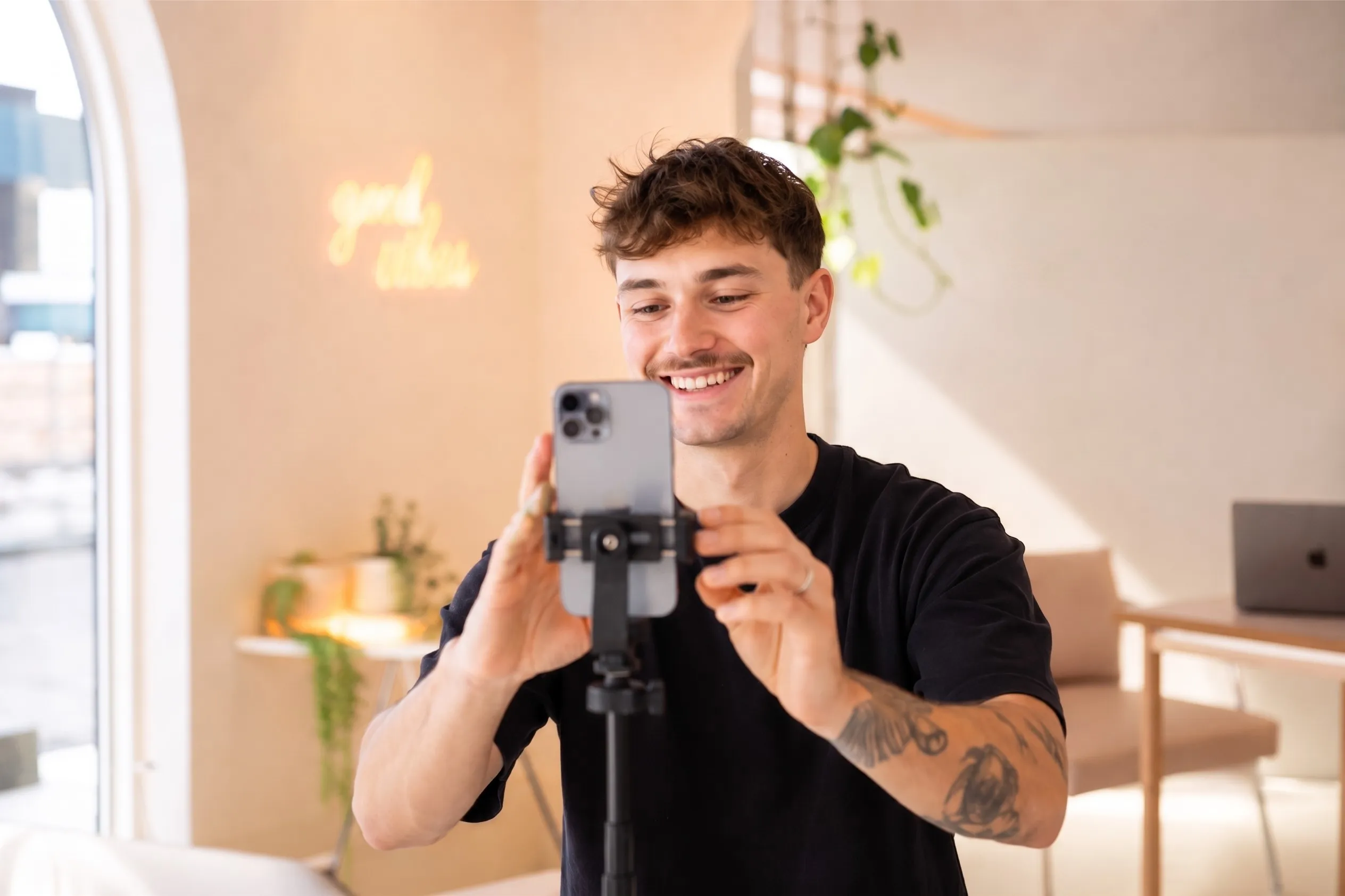 Content creator filming himself on a phone mounted on a tripod in a bright, plant-filled studio