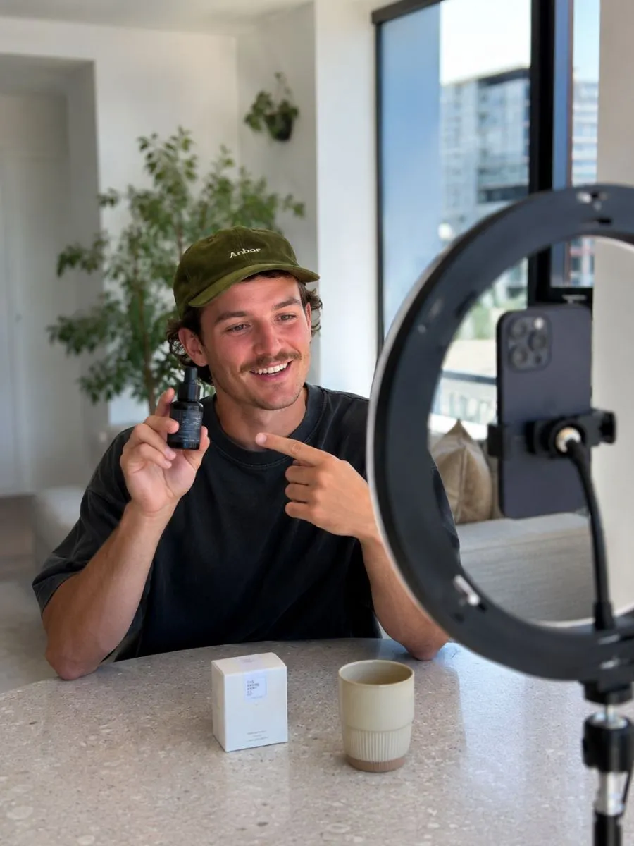 Content creator filming a product review of a skincare bottle on a ring-light phone setup at home