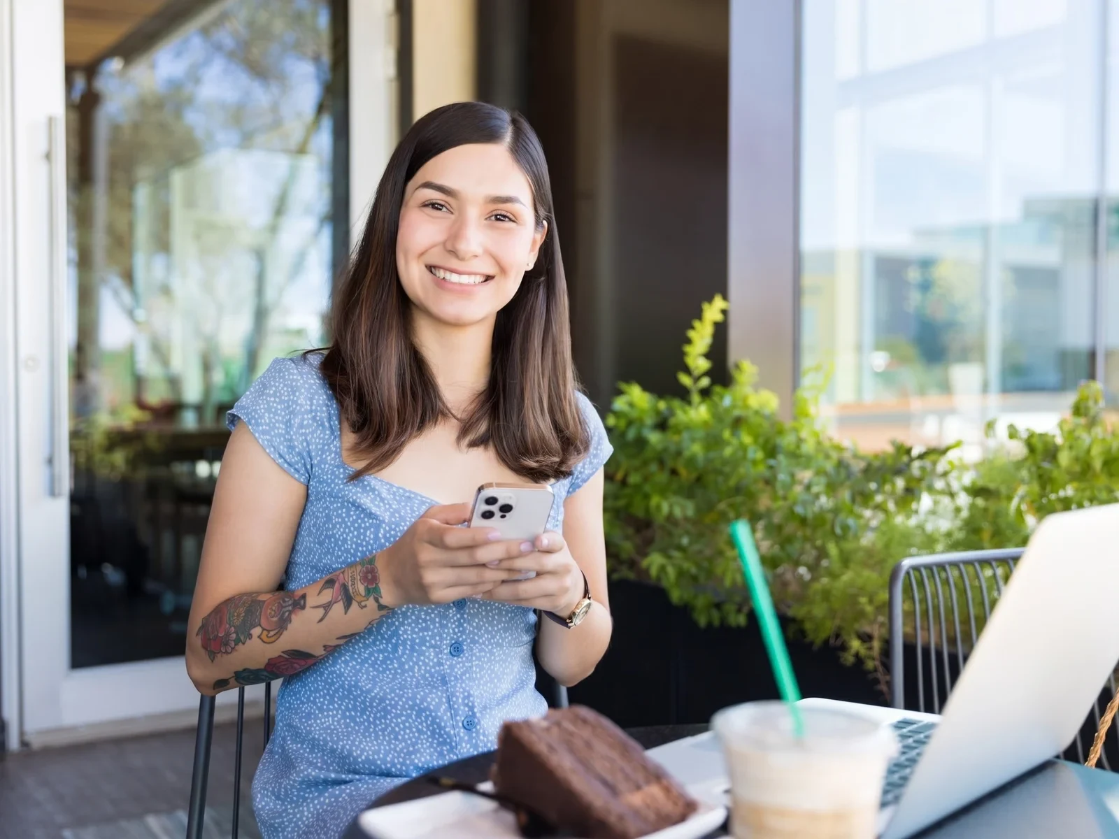 Smiling content creator at an outdoor cafe with her phone, laptop, and iced coffee